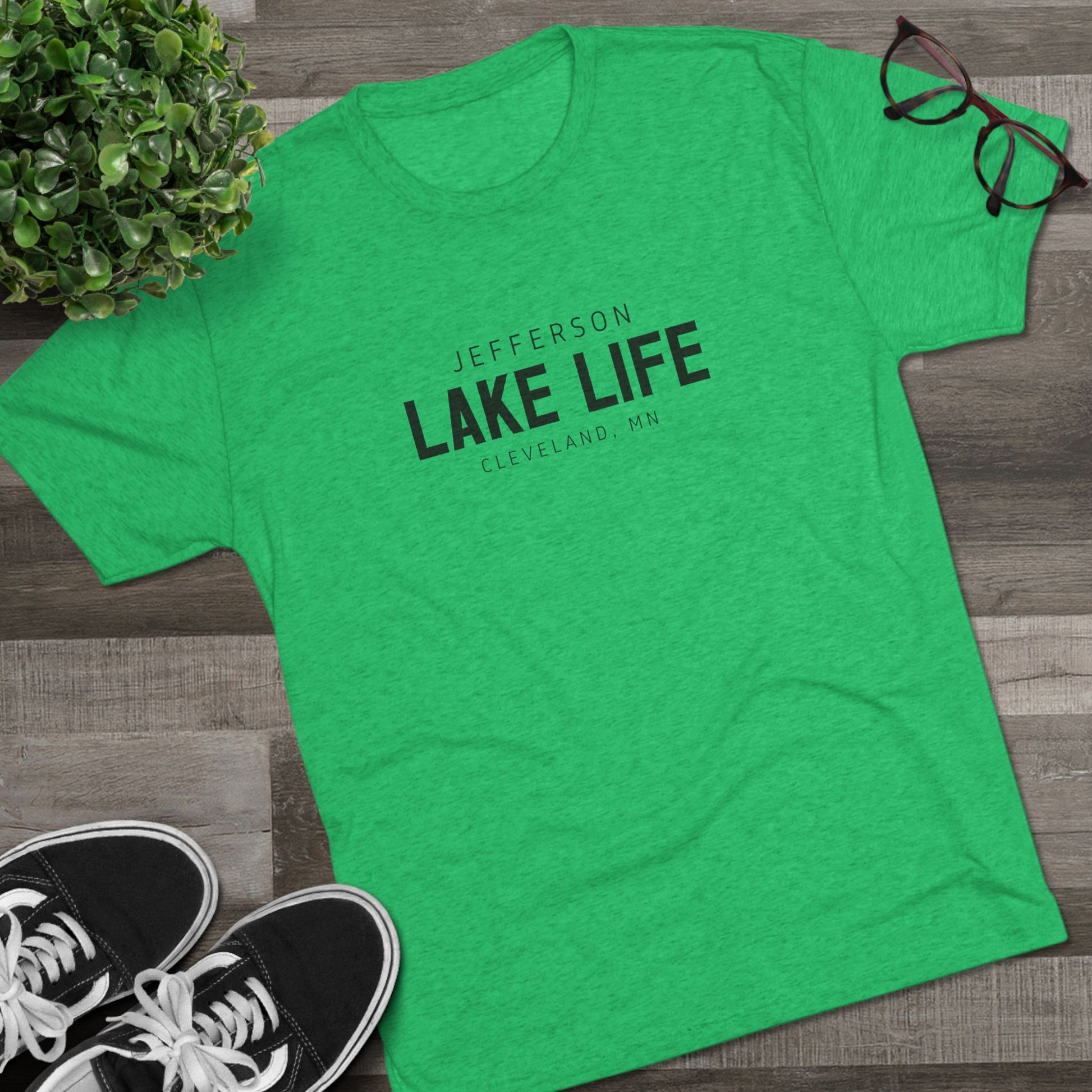 Green t-shirt with 'Jefferson Lake Life Cleveland, MN' text on a wooden surface with shoes and a plant.