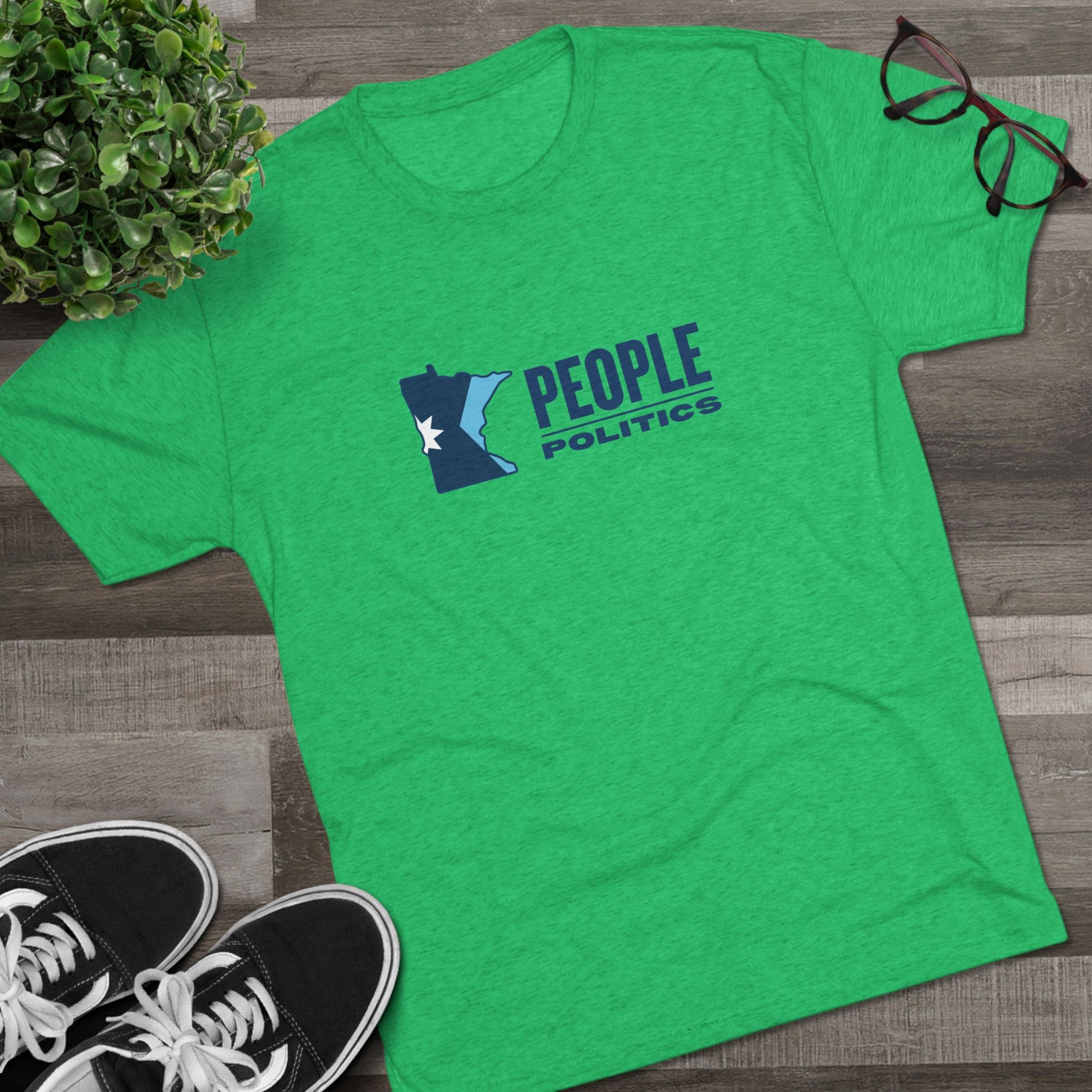 Green t-shirt with 'People Politics' design on a wooden surface with shoes and a plant.