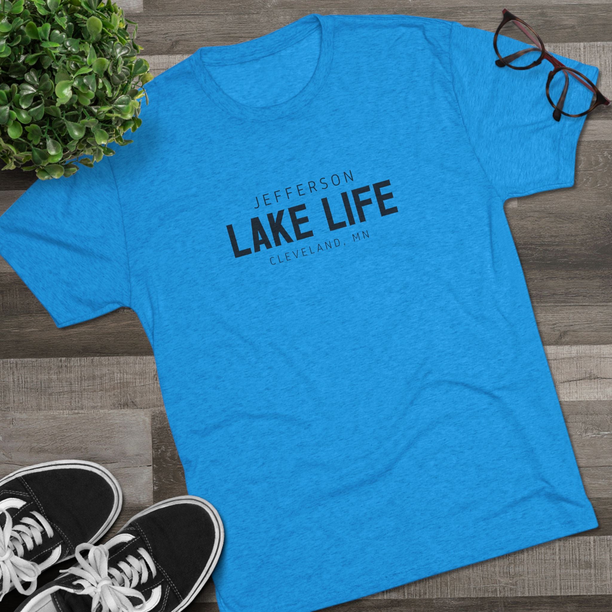 Blue t-shirt with 'Jefferson Lake Life' text on a wooden surface with shoes and a plant.