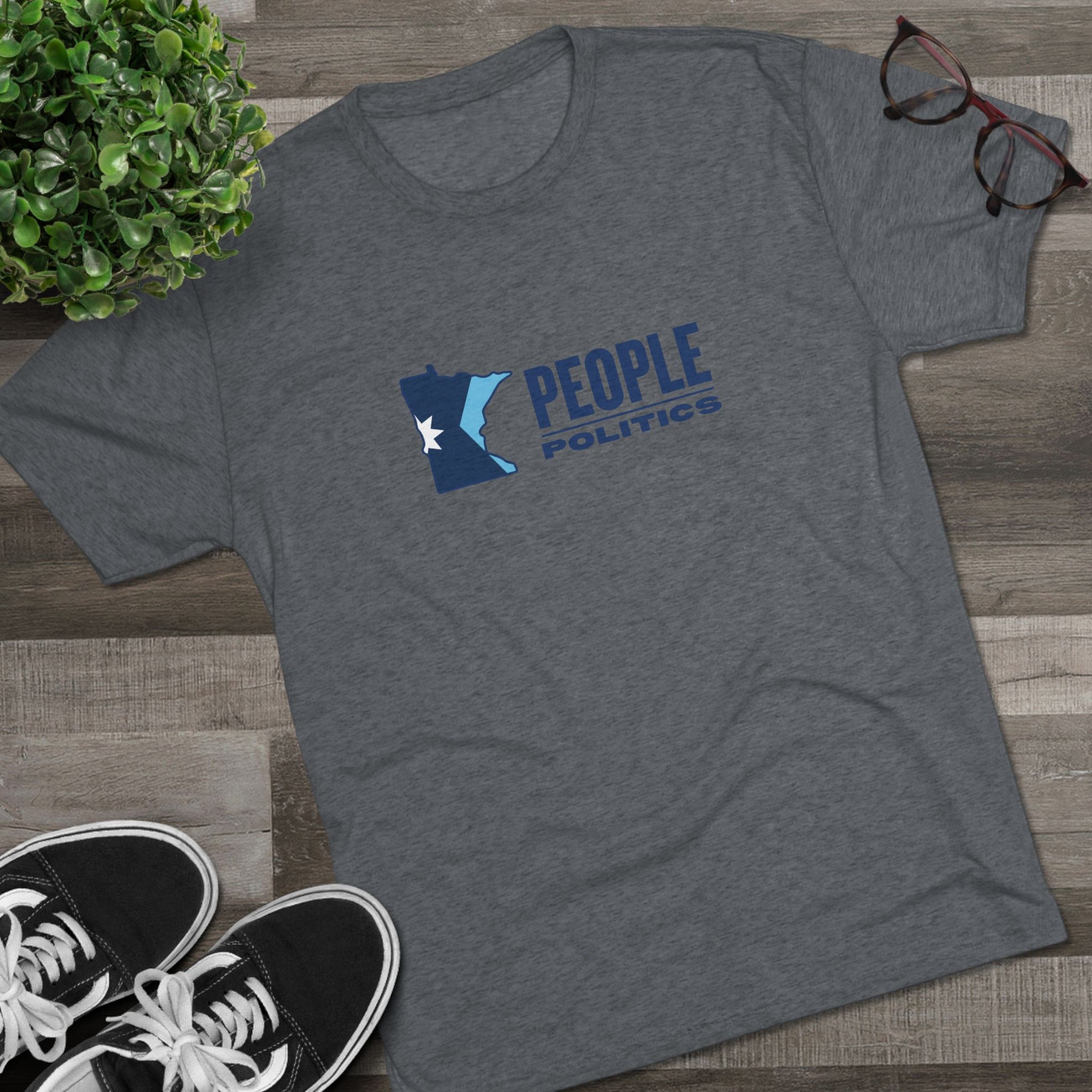 Gray t-shirt with 'People Politics' logo on a wooden surface with sneakers and a plant.