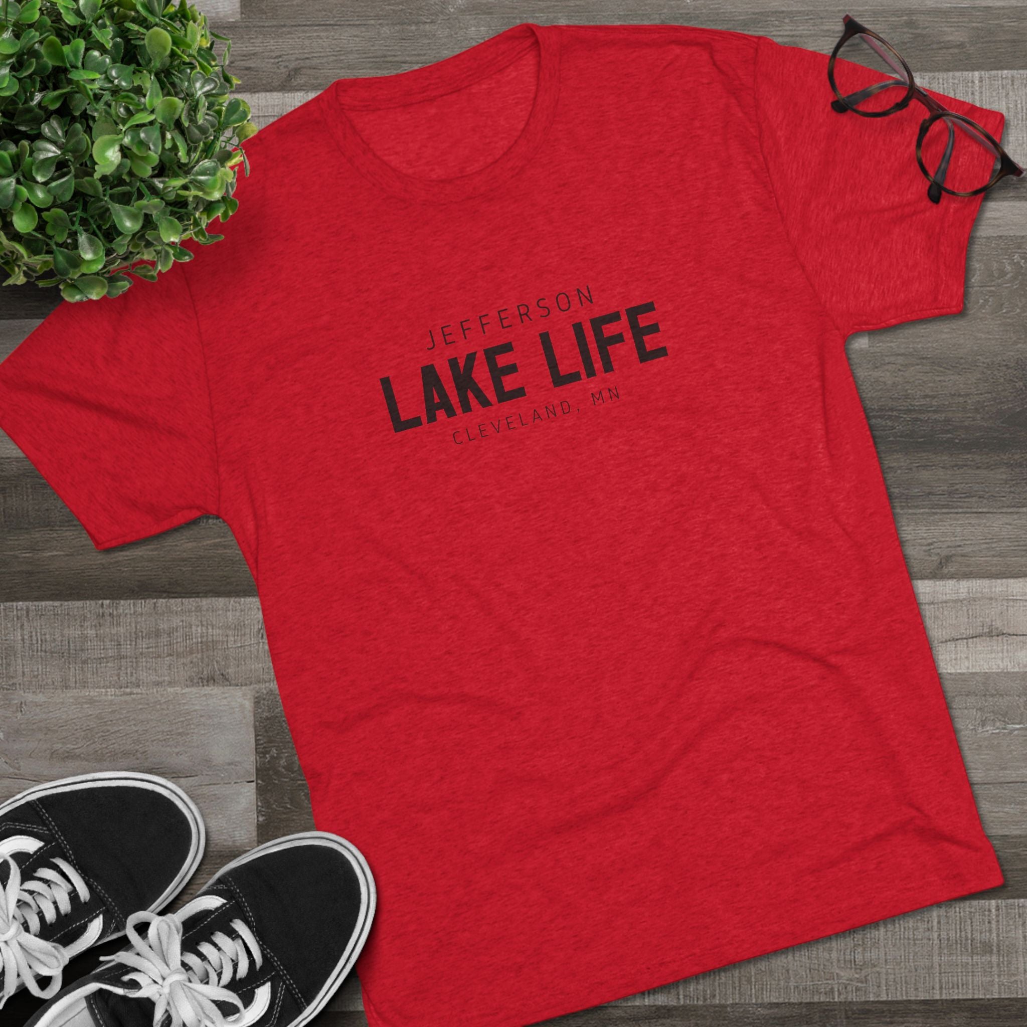 Red t-shirt with 'Jefferson Lake Life' text on a wooden surface with shoes and a plant.