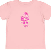 A beige toddler T-shirt with 'Howl Sweet It Is' written in pink, featuring a basketball symbol and a heart at the end.