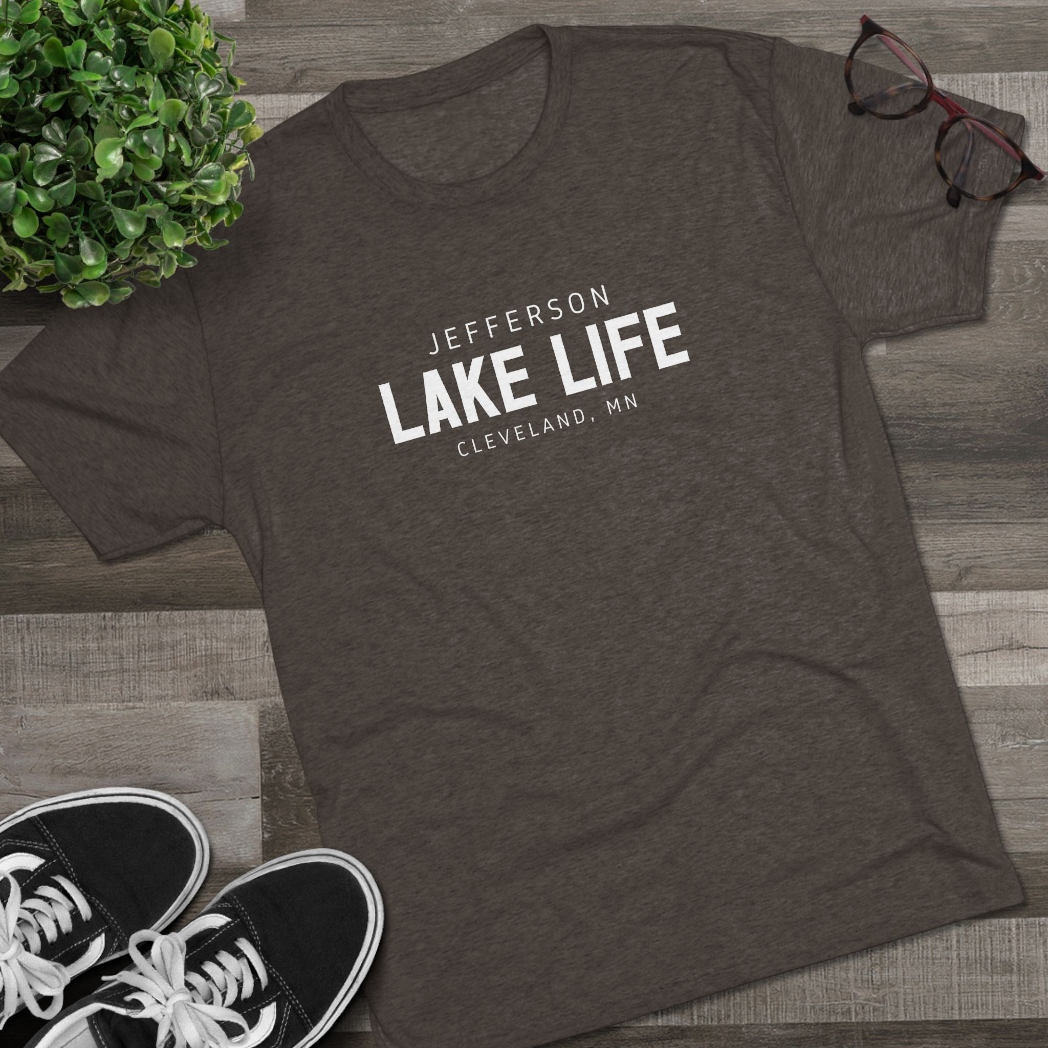 Dark gray t-shirt with 'Jefferson Lake Life, Cleveland, MN' text on a wooden surface with sneakers and a plant.