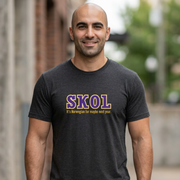 A smiling bald man wearing a dark charcoal gray t-shirt with 'SKOL' printed in purple and gold on the chest, standing outdoors in a blurred urban environment.