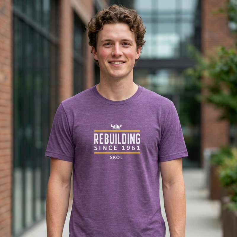 A smiling young man wearing a heather purple t-shirt with a graphic that reads 'REBUILDING SINCE 1981 SKOL' in white, orange, and yellow, standing outdoors in front of a brick building.