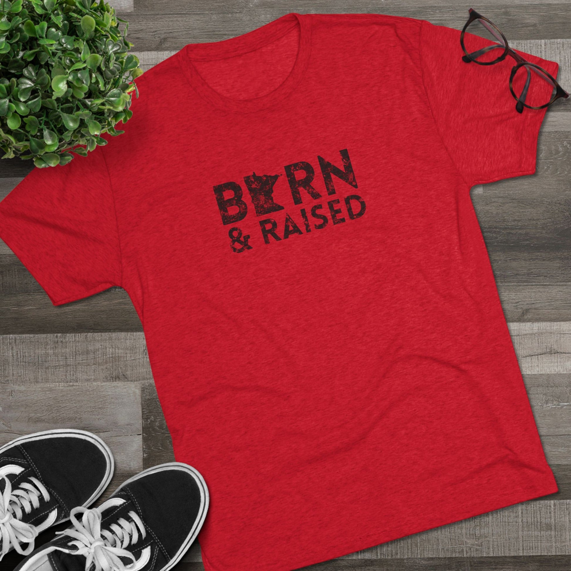 Red t-shirt with 'Born & Raised' text on a wooden surface with sneakers and a plant.