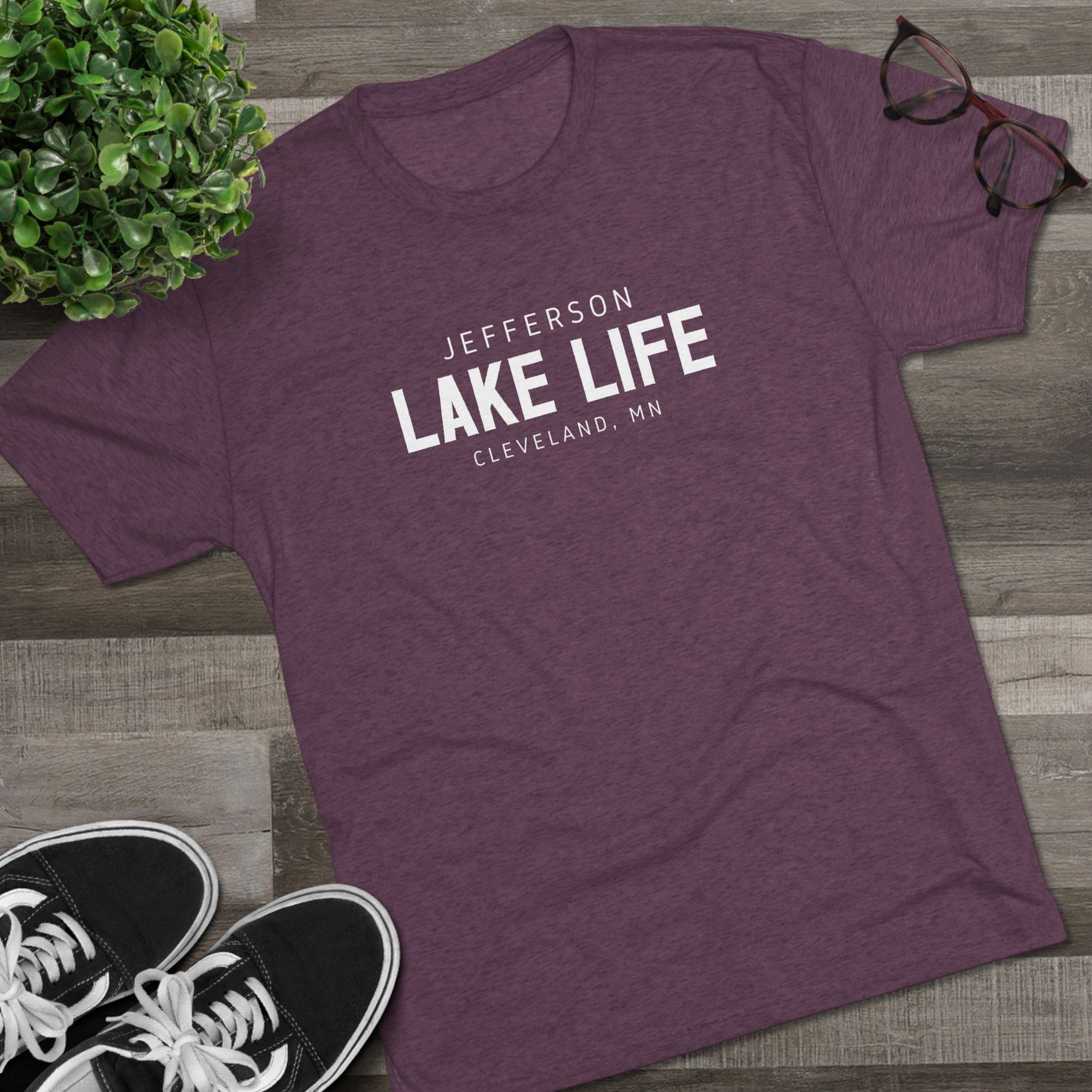 Purple t-shirt with 'Jefferson Lake Life, Cleveland, MN' text on a wooden surface with sneakers and a plant.