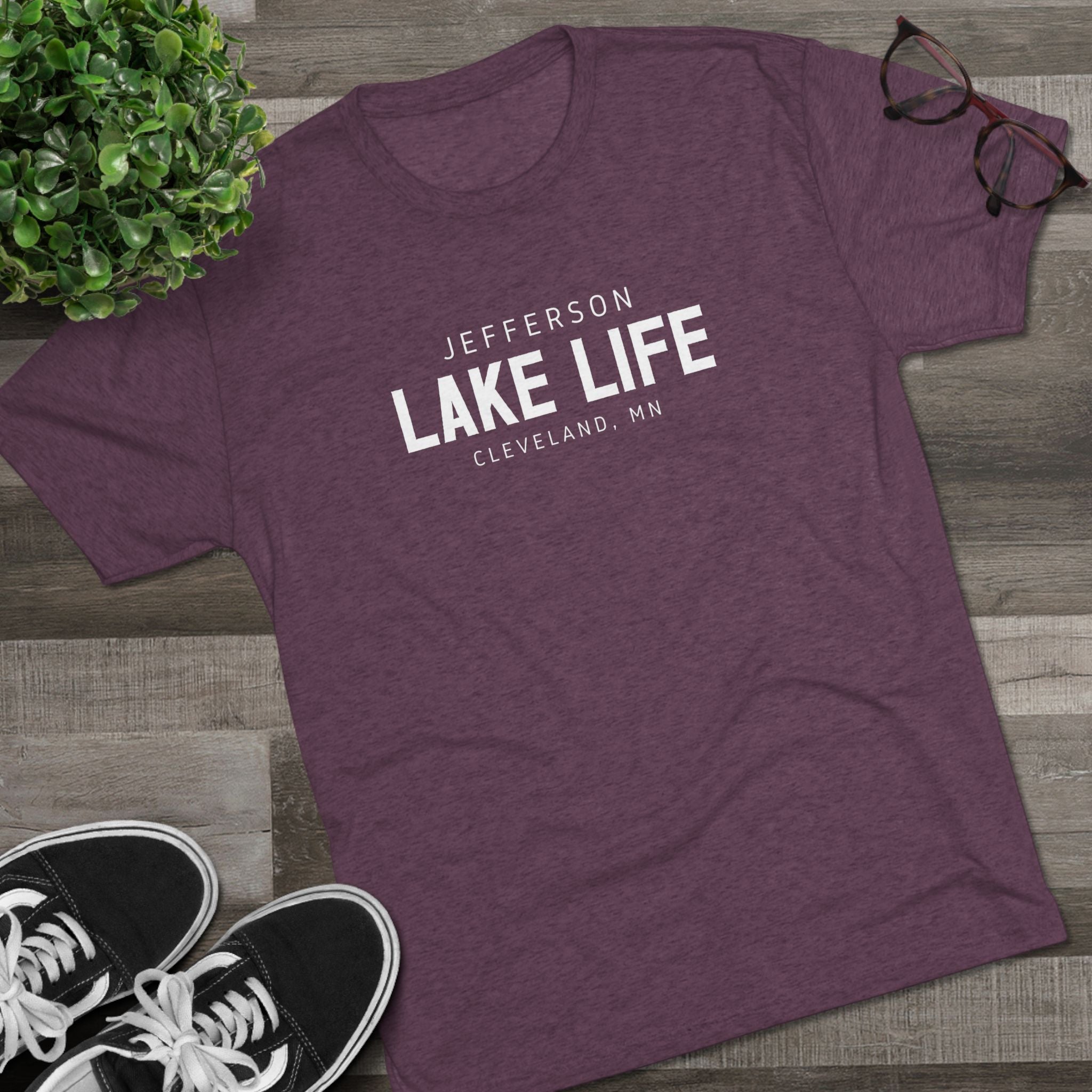 Purple t-shirt with 'Jefferson Lake Life, Cleveland, MN' text on a wooden surface with sneakers and a plant.