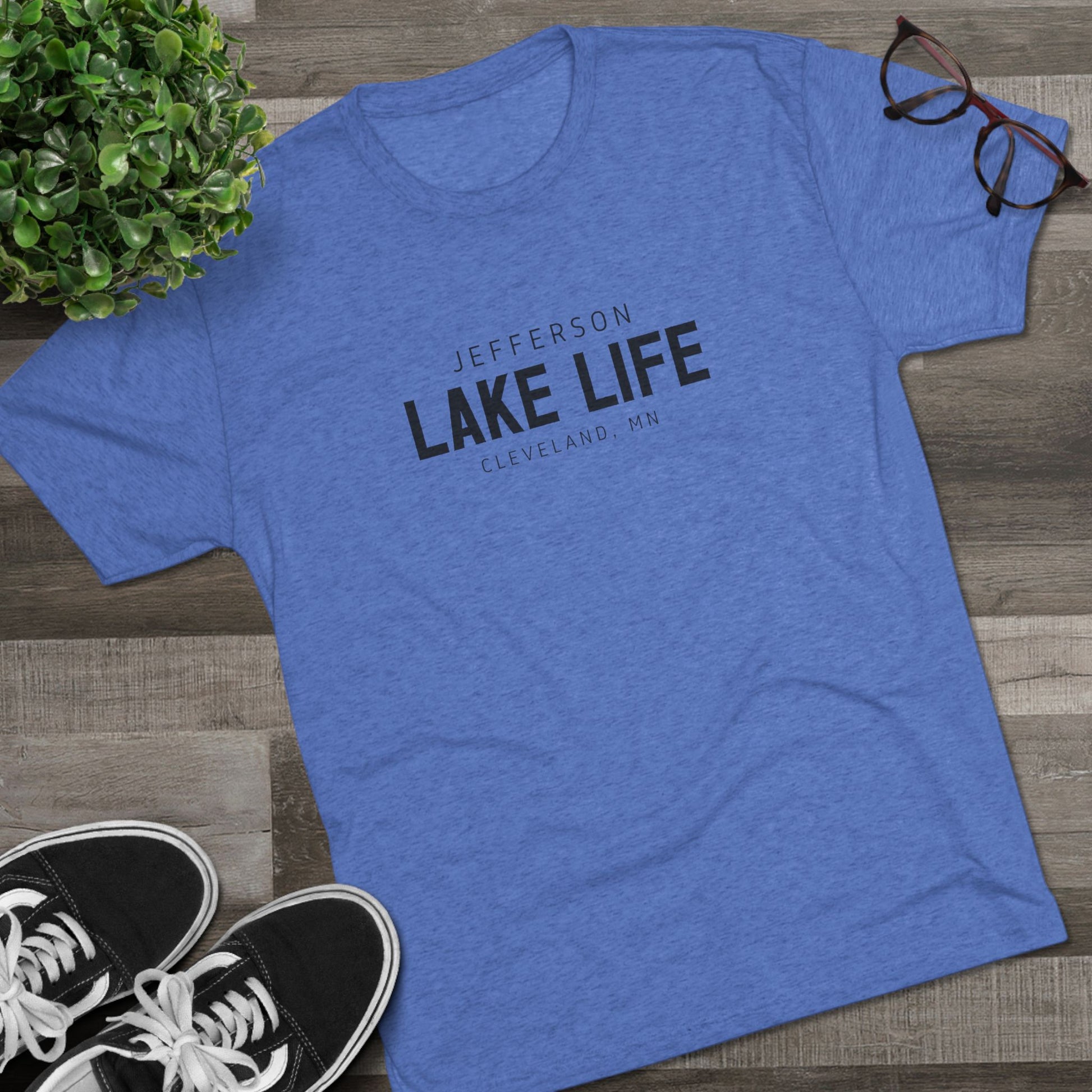 Blue t-shirt with 'Jefferson Lake Life Cleveland, MN' text on a wooden surface with sneakers and a plant.