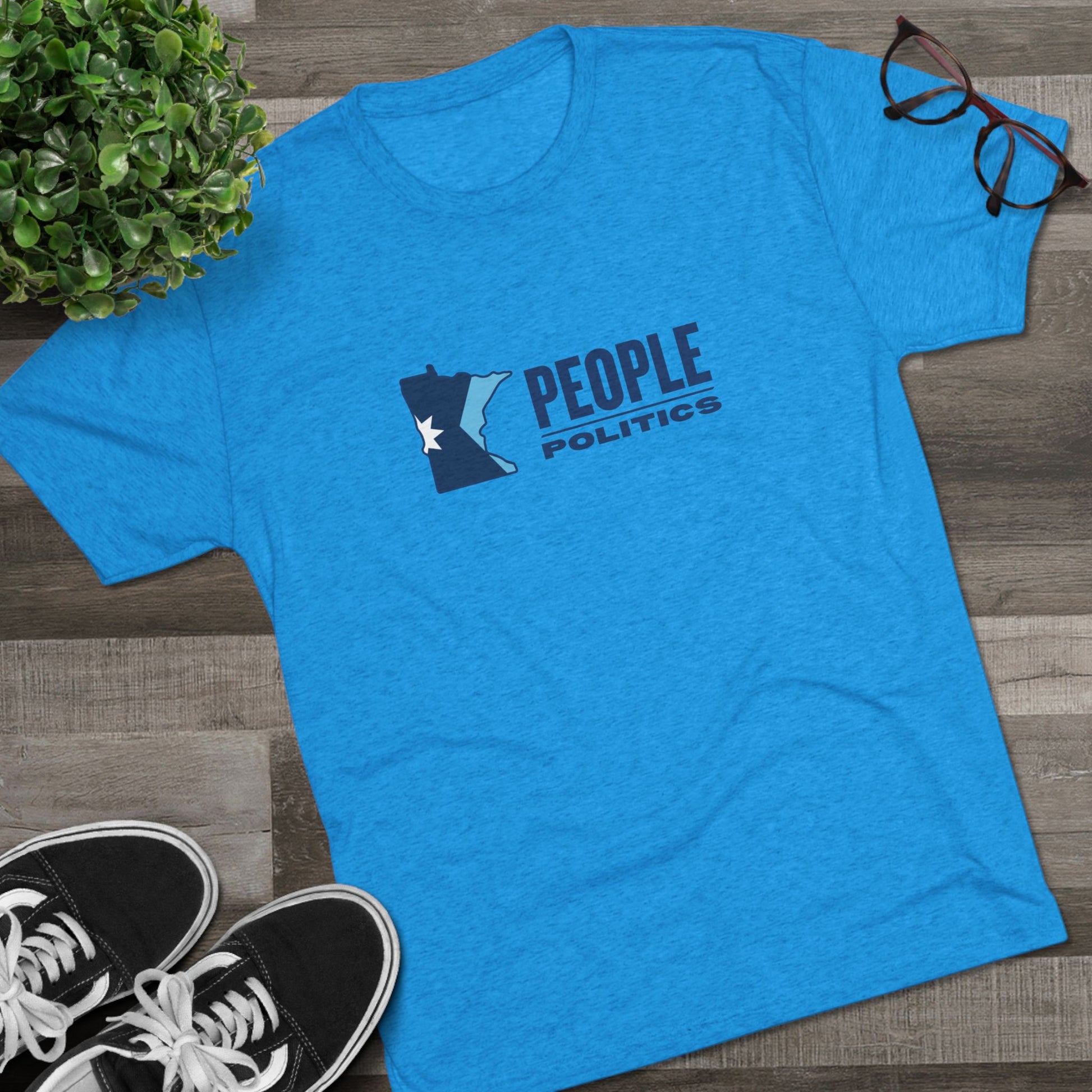 Blue t-shirt with 'People Politics' design on a wooden surface with shoes and a plant.