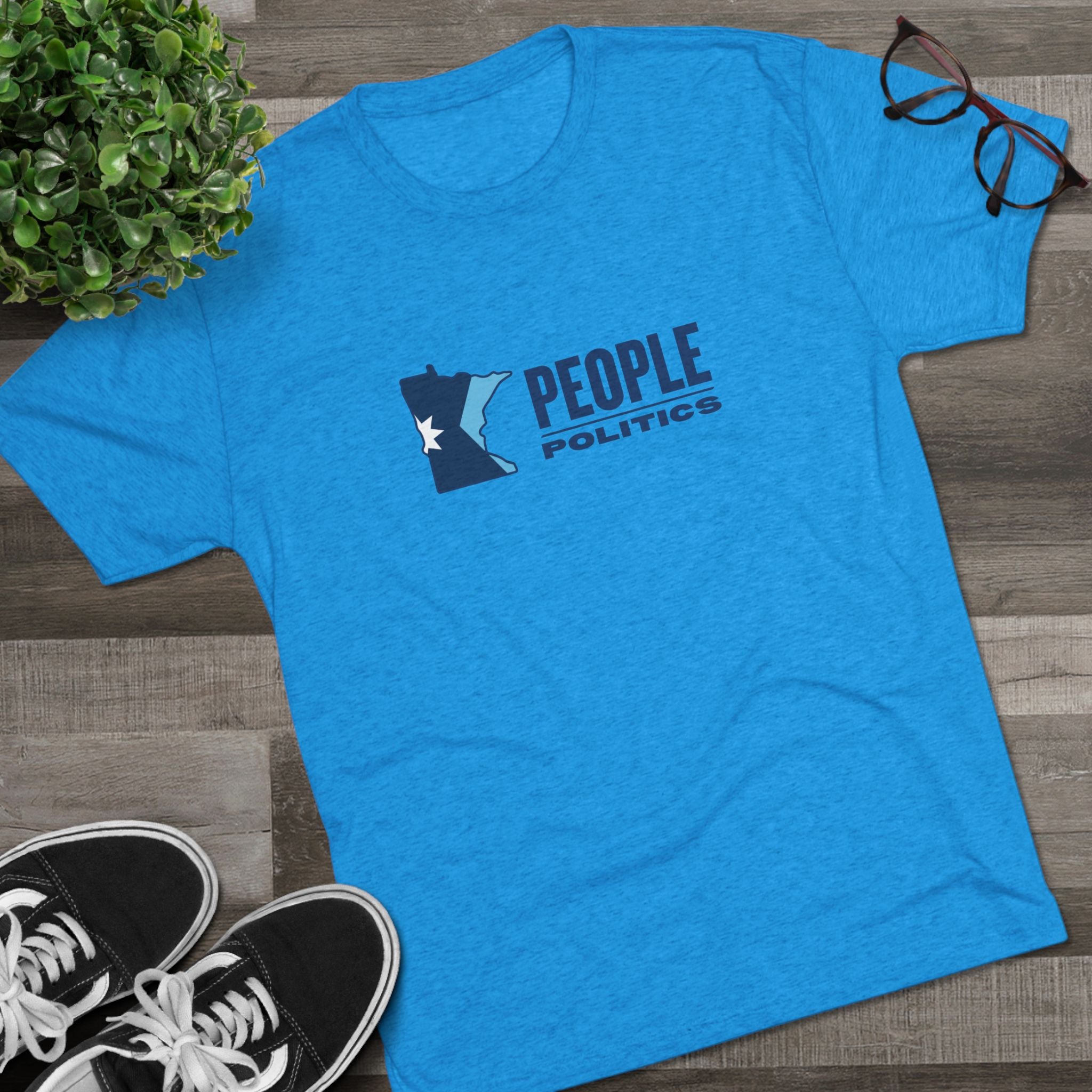 Blue t-shirt with 'People Politics' design on a wooden surface with shoes and a plant.