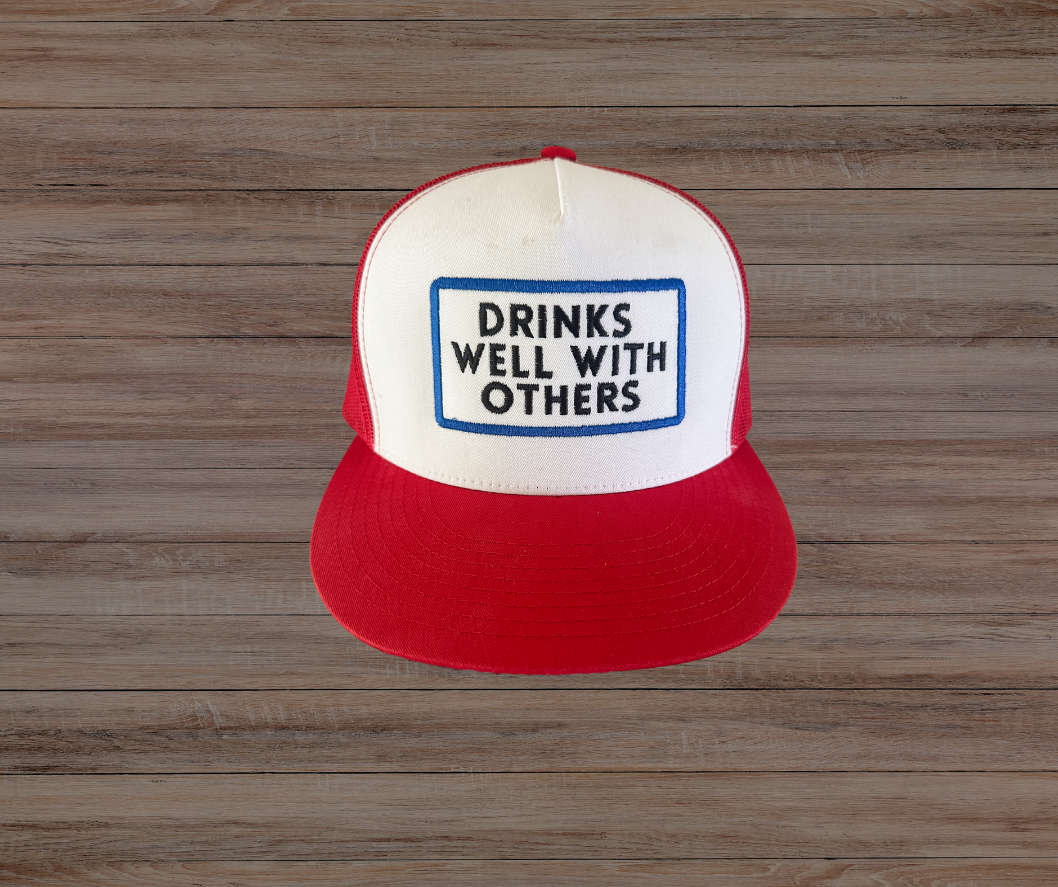 Red and white cap with 'DRINKS WELL WITH OTHERS' text on a wooden background