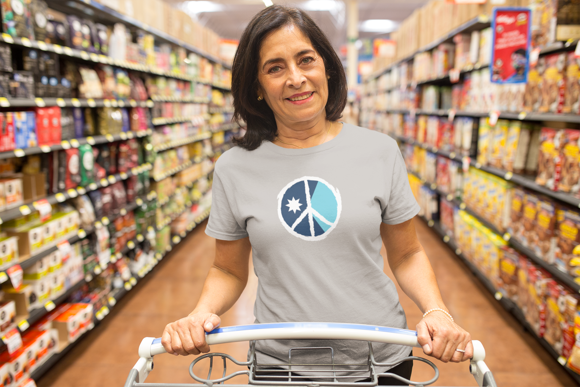 Woman pushing a shopping cart in a grocery store aisle wearing a t-shirt with a peace symbol design.